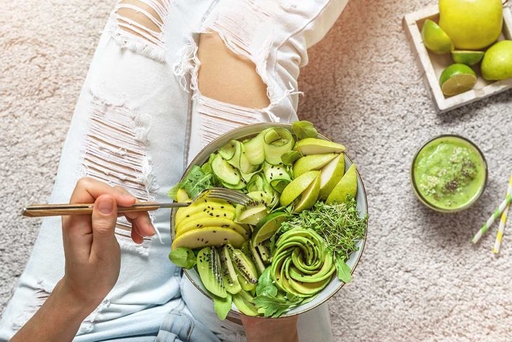 Woman in jeans holding fresh healthy greeen salad with avocado, kiwi, apple, cucumber, pear, greens and sesame on light background. Healthy food, clean eating, Buddha bowl salad, top view, detox